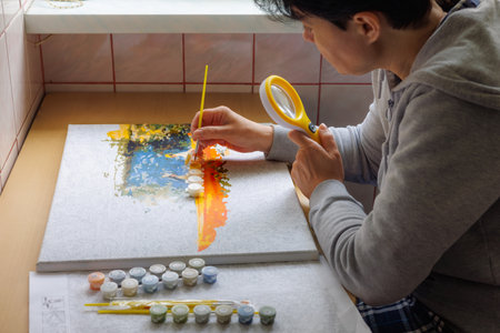 A woman at the window paints a picture by numbers, looking through a magnifying glass. Favorite hobby.の写真素材