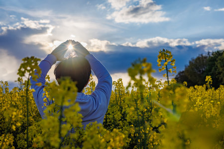 Woman in a rapeseed field looking through her hands at the sun. Sunset sky with clouds.