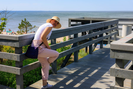 Woman in straw hat bends over and adjusts sneaker on pier with sea in background. Summer beach vacation.の写真素材