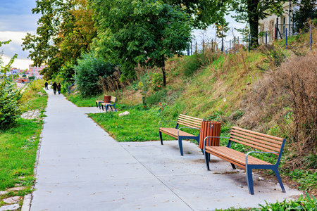 Two wooden benches and a trash bin on a path in a city park.の写真素材