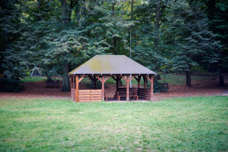 A beautiful wooden gazebo with tables and benches on a green lawn against a backdrop of trees. In...