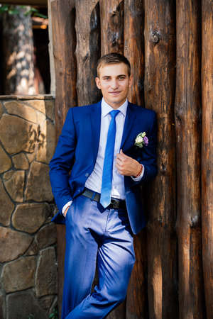 The groom in a suit stands near the wooden wall from logsの写真素材