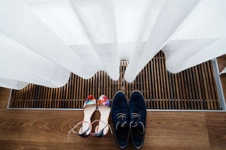 Colorful sandals of the bride and blue shoes of the groom under the curtain at the windowの写真素材