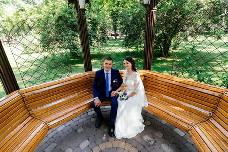 Beautiful young bride and groom sitting on a bench and holding hands.Wedding couple in love at wedding dayの写真素材