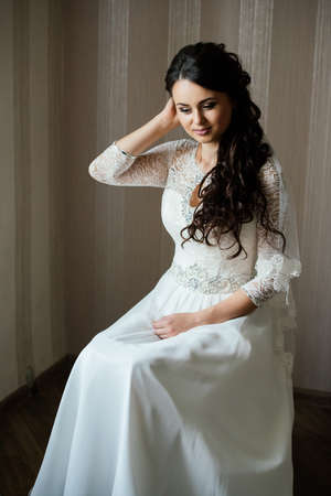 Beautiful young bride in white dress sitting and waiting for the groom indoors. Elegant charming young brunette bride is posing indoors in a wedding dress. Morning, the brideの写真素材