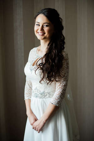Beautiful young smiling bride in white dress waiting for the groom indoors. Elegant charming young brunette bride is posing indoors in a wedding dress. Morning, the brideの写真素材