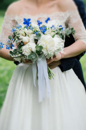 Wedding bouquet of peonies, blue flowers and greenery in the bride's handsの写真素材