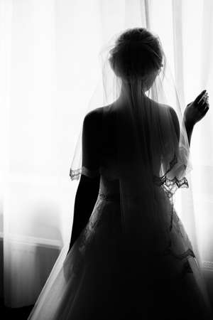 Silhouette of beautiful bride is standing at the window and holding curtains. Morning of the bride. Bride in veil posing in sunlight at window in morning. Getting ready. Black and white photoの写真素材