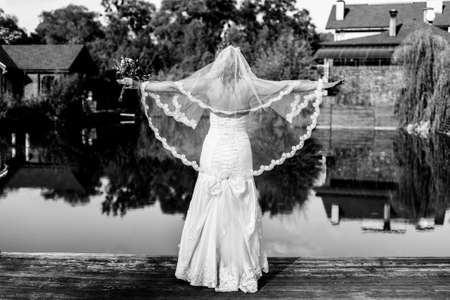 Happy bride in a wedding dress and veil with a bouquet in her hands is standing near the lake with her arms outstretched. Black and white photographyの写真素材