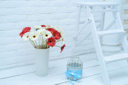 White and red gerberas in white vase near white brick wall and small decorative metallic cageの写真素材