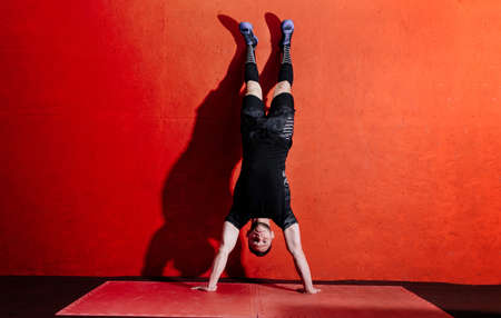 Athlete doing push ups on his hands while standing upside down near red wall. Full body length...