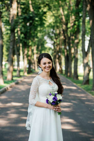 Beautiful young smiling brunette bride in wedding dress with bouquet of flowers in hands outdoors on the background of green leavesの写真素材