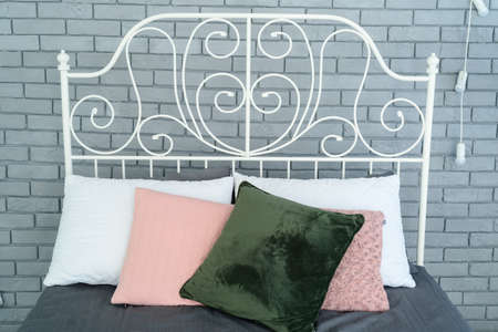 Colored pillows on white metal frame bed with decorative metal headboard near grey brick wallの写真素材