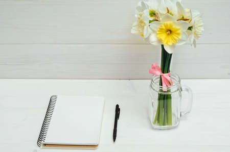 Yellow bouquet of narcissus in vase on white wooden background. Blank card flat lay. Top view on table with narcissus, empty diary and pencil, free spaceの写真素材