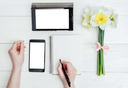 Top view on table with narcissus, empty diary, tablet, phone and girl's hands with pen, free space. Girl's hands writing in diaryの写真素材
