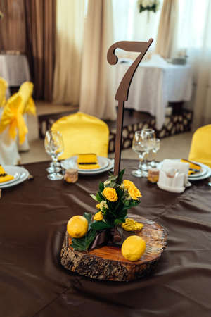 Wedding flower arrangement of yellow roses, greenery and lemons on stump under table number card on wedding dinner table in blur. Number of guest table at wedding banquetの写真素材