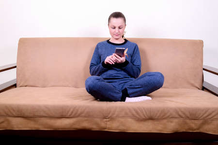 Beautiful young woman sitting on a sofa, using a smartphone and texting. Relaxing on the couch at home. Technology and communication conceptの写真素材