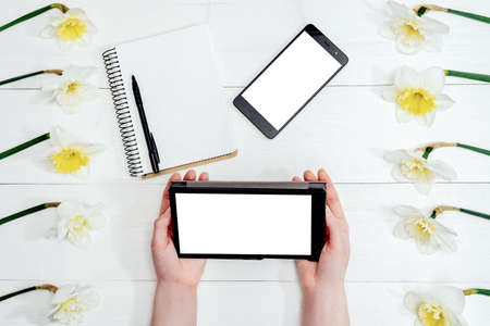 Top view on table with narcissus, empty diary, phone and girl's hands with tablet, free space. Girl's hands writing in diaryの写真素材