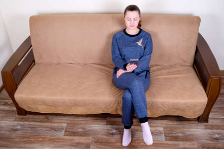 Beautiful young woman sitting on a sofa, using a smartphone and texting. Relaxing on the couch at home. Technology and communication conceptの写真素材