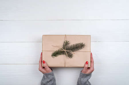 Woman with red manicure holding Christmas present  wrapped in kraft paper on white wooden table...