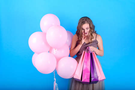 Happy young woman looking at shopping bags near pink balloons over blue background in black friday holiday, free space. Beauty, fashion, seasonal sale concept. Girl on blue background with copy spaceの写真素材
