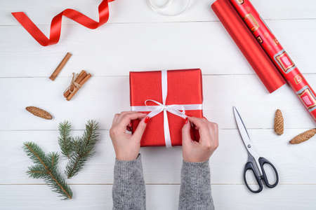 Christmas gift wrapping background. Female hands packaging christmas present with white satin ribbon, top view. Winter holidays concept, flat lay. Woman holding red Christmas giftの写真素材