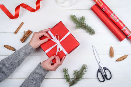 Christmas gift wrapping background. Female hands packaging christmas present with white satin ribbon, top view. Winter holidays concept, flat lay. Woman holding red Christmas giftの写真素材