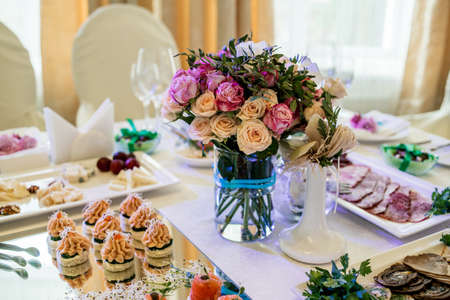 Beautiful wedding bouquet of cream and pink roses in glass vase on dinner table, selective focus. Table setting at a luxury wedding reception. Flowers on the tableの写真素材