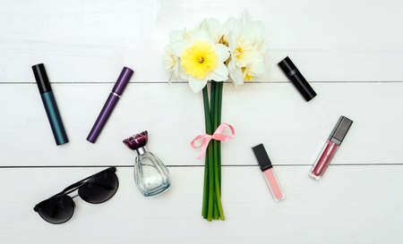 Top view on white wooden background with Ñosmetics: mascara, lip gloss, lipstick and perfume around bouquet of narcissus. Liquid Lipstickの写真素材