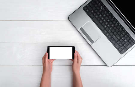 Top view on white wooden table with open blank laptop computer and girl's hands with cell phone, free space. Mobile phone with white screen, copy space の写真素材