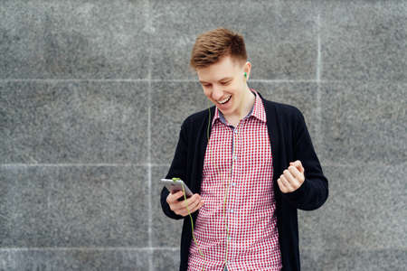 Cheerful happy young man in plaid shirt and jacket listening to music with headphones from cell phone and singing outdoors. Student with smart phone and headphonesの写真素材