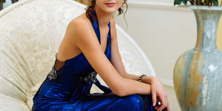 Young beautiful slim brunette girl in long blue dress with open shoulders and decollete sitting with crossed hands on chair in the roomの写真素材