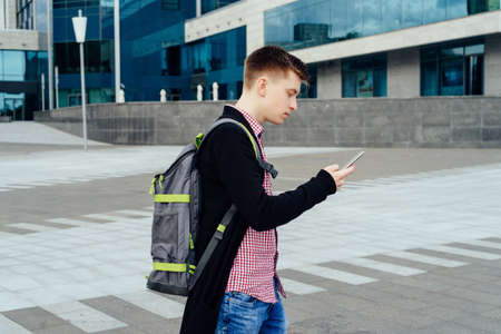 Stylish young man in plaid shirt with backpack walking in the city and using smartphone outdoors. Student with smartphone walking の写真素材