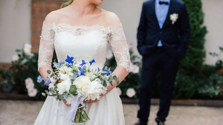 Beautiful bride in wedding dress with wedding bouquet of peonies, blue flowers and greenery with groom on background in blur outdoorsの写真素材