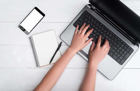 Top view on white wooden table with open blank laptop computer, cell phone, empty diary and girl's hands typing on keyboard, free space. Mobile phone with white screen, copy space の写真素材