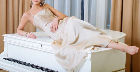 Young beautiful slim brunette girl in long beige dress with open shoulders lying on white piano in the roomの写真素材