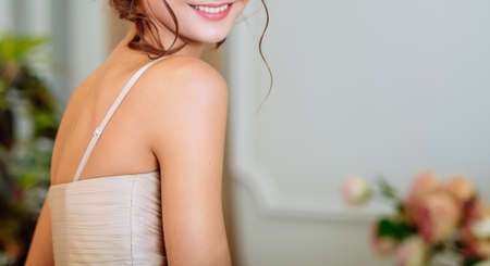 Young beautiful slim smiling brunette girl in beige dress with open shoulders in the room indoorsの写真素材