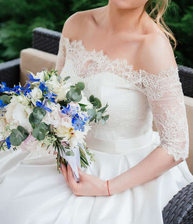 Beautiful bride in wedding dress with wedding bouquet of peonies, blue flowers and greenery sitting on couch outdoorsの写真素材