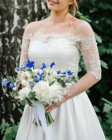 Beautiful bride in wedding dress with wedding bouquet of peonies, blue flowers and greenery standing near birch outdoorsの写真素材
