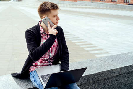 Stylish young man in plaid shirt sitting in the city and talking on cell phone with laptop computer on knees outdoors. Student with mobile phone and laptopの写真素材