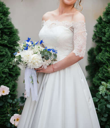 Beautiful bride in wedding dress with wedding bouquet of peonies, blue flowers and greenery outdoorsの写真素材