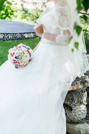 Beautiful bride in white lace wedding dress with wedding bouquet of cream and pink roses, purple flowers sitting on bench at stone table outdoors in sunny dayの写真素材