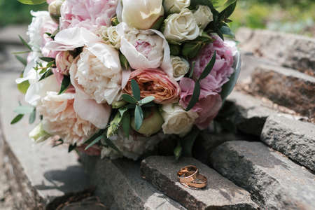 Two golden wedding rings on stone near   beautiful wedding bouquet of white and pink peony and roses outdoors, close upの写真素材