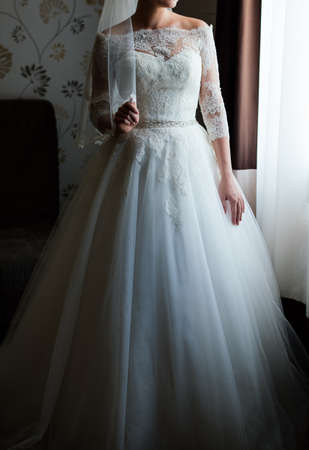 Beautiful young bride in white dress waiting for the groom indoors. Elegant charming young brunette bride posing indoors in wedding dress. Morning, the brideの写真素材