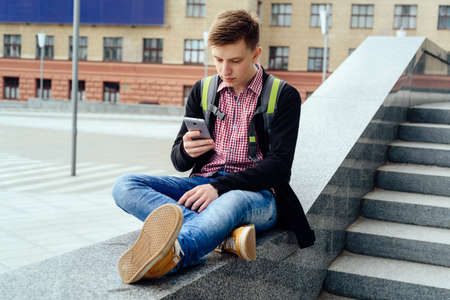Happy handsome young man with backpack sitting on stone near stairs and using smart phone in the city outdoors, full body.  Student rests after college outdoorsの写真素材