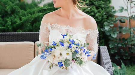 Beautiful bride in wedding dress with wedding bouquet of peonies, blue flowers and greenery sitting on couch outdoorsの写真素材
