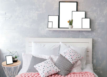 White and colored pillows on woodenl bed with white headboard near grey wall. Shelf with photo frames with blank empty white cards above the bed with set of pillows and pink merino wool blanket near windowの写真素材