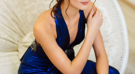 Young beautiful slim brunette girl in long blue dress with open shoulders and decollete sitting with hand on shoulder on chair in the roomの写真素材