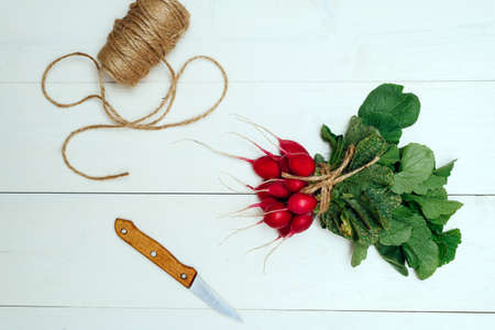 Top view of bunch of fresh organic red radishes with tops and green leaves, knife and rope on white wooden background. Vegetable backgroundの写真素材