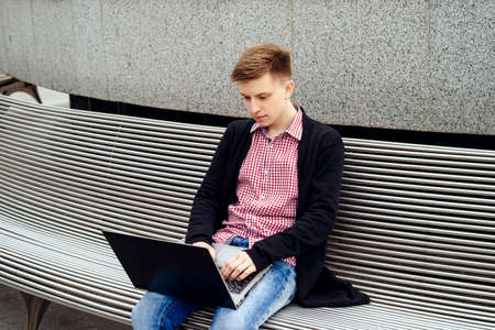 Stylish young man in jacket and jeans sitting on the bench and typing on laptop computer outdoors. Technology and communication concept の写真素材